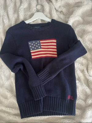 Mörkblå stickad tröja Polo Ralph Lauren - Mörkblå stickad tröja från Polo Ralph Lauren med amerikansk flagga framtill och RL-broderi vid nederkanten. Tröjan har rund halsringning och ribbstickade muddar vid ärmslut och nederkant. Perfekt för dig som gillar klassisk preppy stil.
