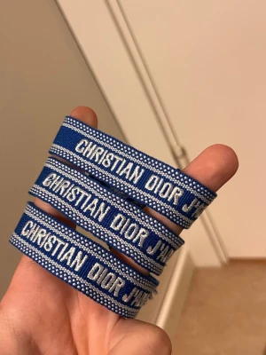 Blått armband från Christian Dior - Snyggt blått armband från Christian Dior med vit broderad logga. Armbandet är gjort i textil och har en trendig, sportig look med tydlig branding. Perfekt accessoar för att lyfta din outfit.
