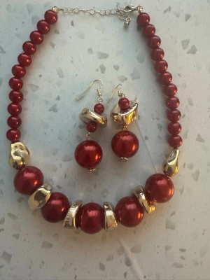 Rött pärlset med halsband och örhängen - Snyggt set med halsband och matchande örhängen i glansiga röda pärlor och guldfärgade detaljer. Halsbandet har stora och små pärlor i plast och metall, och örhängena har krokar för enkel användning. Perfekt för att ge din outfit en färgklick.