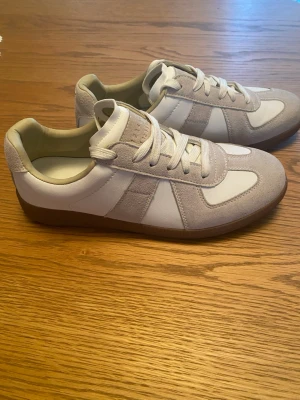 Margiela skor 38/39 - Snygga GAT sneakers i vitt och beige med ovandel i mocka och släta partier i läder. Klassisk låg modell med bruna sulor och vita skosnören. Perfekta för en clean och stilren look. Ej säker på äktheten därav priset.