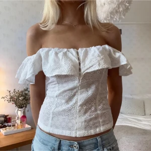 Vit offshoulder topp - Säljer en superfin vit offshoulder blus med volang och spetsdetaljer. Toppen är figurnära och har ett romantiskt uttryck med broderade blommor och luftig känsla. Perfekt till jeans eller kjol för en trendig look.