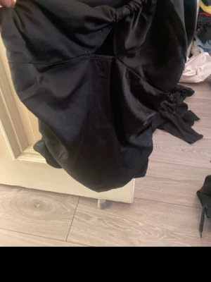Svart satin linne med rosett - Snygg svart kort klänning från NA-KD i glansigt satinmaterial. Klänningen har tunna axelband och en dekorativ rosett framtill. Perfekt för dig som vill ha en stilren och trendig look.