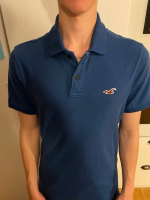 Blå pikétröja från Hollister - Snygg blå pikétröja från Hollister med klassisk krage och två knappar framtill. Tröjan har det ikoniska Hollister-logotypen broderad på bröstet och är tillverkad i ett stretchigt material för extra komfort.