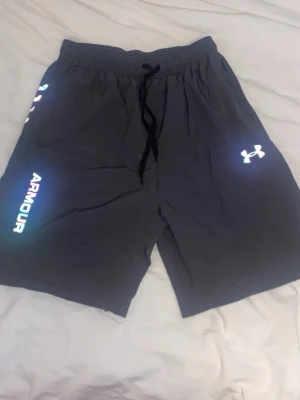 Svarta shorts från Under Armour - Snygga svarta shorts från med reflexdetaljer och logga på ena benet. Shortsen har elastisk midja med snörning och är tillverkade i ett lätt och bekvämt material, perfekta för träning eller chill.