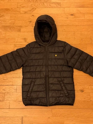 Svart pufferjacka från Lyle & Scott - Säljer en svart pufferjacka från Lyle & Scott med huva och dragkedja framtill. Jackan har två snedställda fickor och det klassiska gula logomärket på bröstet, med två innerfickor på vardera sida. Perfekt för kalla/blåsiga dagar. Pris kan diskuteras!😊