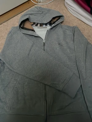 Grå zip hoodie från Burberry - Snygg grå hoodie med dragkedja från Burberry. Klassisk huva med det ikoniska rutmönstret på insidan och diskret broderad logga på bröstet. Tillverkad i mjukt bomullsmaterial och har ribbade muddar vid ärmslut och nederkant. Perfekt för en chill och stilren look.