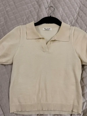 Beige stickad polotopp Paris Et Moi - Beiges stickad polotopp från Paris Et Moi med korta ärmar och klassisk krage. Toppen har en v-ringad öppning framtill och är ribbad vid ärmslut och nederkant. Perfekt för en stilren och enkel look. Är dock använd och råkat få sminkfläck på