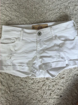 Vita jeans shorts från Hollister - Säljer ett par vita shorts från Hollister med låg midja och slitna detaljer framtill. Klassisk femficksmodell med knapp och dragkedja. Perfekta för varma dagar och enkel att matcha med allt. I storlek W23. Ge prisförslag!!