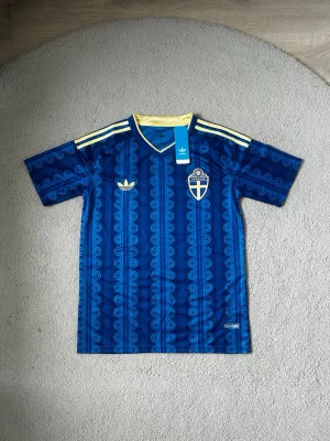 Blå Sverige tröja från Adidas - Säljer en blå Sverige tröja från Adidas med gul logga och emblem på bröstet. Tröjan har gula detaljer på axlarna och ett mönster i olika blå nyanser. Tillverkad i ett lätt och svalt material, perfekt för fotboll eller träning. Den är i bästa kvalité och helt ny, därför är den 10 av 10 i skick.Väldigt fin och bra för sommaren och för träningen. Fraktar så fort du har köpt🚚📦Aktiv 24/7✅ Hör av dig via frågor⁉️