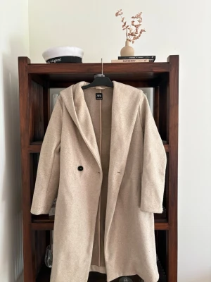 Beige kappa med huva från Zara - Stilren beige kappa från Zara med stor huva och enkelknäppning. Kappan har rak passform, långa ärmar och är tillverkad i ett mjukt, ull-liknande material. Perfekt för dig som gillar minimalistisk och trendig stil.