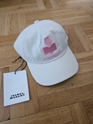 Vit keps från Isabel Marant - Snygg vit keps från Isabel Marant med broderad rosa logga och text framtill. Klassisk böjd skärm och justerbar baksida. Tillverkad i ett tåligt bomullstyg som ger en clean och trendig look.