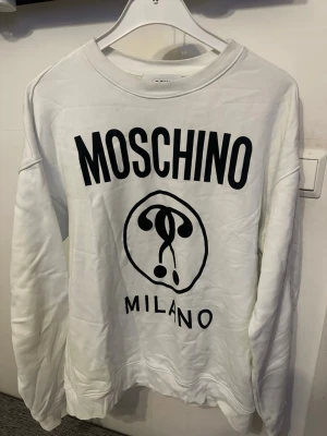 Vit sweatshirt från Moschino - Snygg vit sweatshirt från Moschino med svart tryck framtill där det står 'MOSCHINO MILANO' och en stor logga. Tröjan har rund halsringning och långa ärmar. Perfekt för dig som gillar streetwear och vill ha en statement-piece i garderoben. Storlek 50 eu som motsvarar M/L