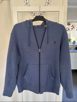 Blå zip hoodie från Polo Ralph Lauren - Snygg blå hoodie med dragkedja från Polo Ralph Lauren. Klassisk design med huva och dragsnören, känguruficka framtill och diskret logga broderad på bröstet. Tillverkad i mjukt bomullsmaterial som känns skönt mot huden. Hoodien är använd max 5 gånger!