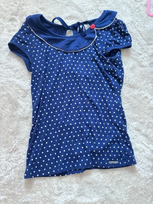 Blå prickig topp med krage och knyt - Söt blå topp från Pussy Deluxe med vita prickar, dekorativ sjömanskrage med vit kant, röd knappdetalj och knyt i nacken. Kortärmad och i mjuk bomull, perfekt för en lekfull och trendig look.