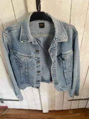 Ljusblå jeansjacka från Lee - Klassisk ljusblå jeansjacka från Lee med två bröstfickor och metallknappar framtill. Jackan har en avslappnad passform och snygga söm-detaljer. Perfekt att slänga över en hoodie eller t-shirt för en cool och tidlös look.