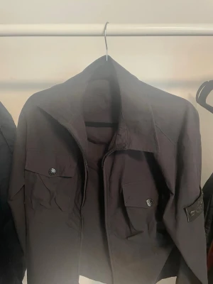 Overshirt Stone Island - Snygg svart jacka från Stone Island med klassiska ghost badge på ärmen. Jackan har två bröstfickor med knappar och dragkedja framtill. Perfekt för dig som gillar streetwear och vill ha en stilren look. Endast testad, skickar inom 24h