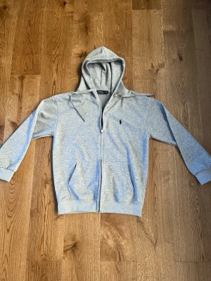 Grå hoodie från Polo Ralph Lauren - Snygg grå zip-hoodie från Polo Ralph Lauren med klassisk huva och dragsko. Tröjan har två fickor framtill och den ikoniska lilla loggan broderad på bröstet. Tillverkad i mjuk bomullsblandning som är perfekt för chill dagar. Storlek medium till large