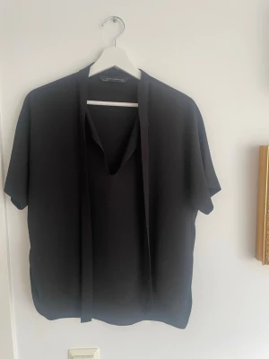 Svart blus med knytband Zara Woman - Stilren svart blus från Zara Woman med korta ärmar och dekorativt knytband vid halsen. Blusen har en rak passform och är tillverkad i ett mjukt, lätt material som ger ett snyggt fall. Perfekt för en trendig, enkel och elegant look🥰