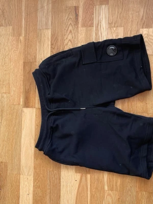 Svarta shorts från C.P. Company - Svarta shorts från C.P. Company med en ikonisk linsdetalj på benfickan. Mjukt material och elastisk midja för skön passform. Perfekta för chill dagar eller streetwear-stil. Klassisk design med fickor och ribbade detaljer. Storlek M passar S också. Priset kan diskuteras för seriösa köpare. 