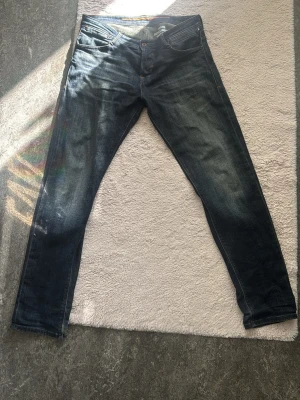 Jack and Jones jeans - Säljer ett par mörkblå slim fit jeans med snygga slitningar och klassisk femficksdesign. Jeansen har kontrastsömmar och en något tvättad look som ger en cool vibe. Perfekta till en avslappnad stil och passar till det mesta i garderoben.