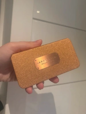 Anastasia Beverly Hills rougepalett - Säljer en snygg rougepalett från Anastasia Beverly Hills i en glittrig guldig förpackning. Paletten innehåller tre nyanser: en persikorosa, en skimrande guld och en varm brun ton. Perfekt för att skapa glow och färg på kinderna.
