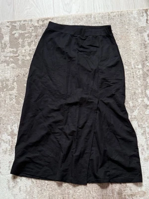 Svart kjol med slits - Stilren svart knälång kjol med diskret slits framtill. Kjolen har en rak passform och bred linning i midjan. Perfekt att matcha med både sneakers och boots för en trendig look.