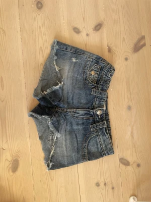 Blå jeansshorts med slitningar - Säljer ett par blå jeansshorts med råa kanter och slitna detaljer. Shortsen har klassiska fem fickor, bälteshällor och knappstängning. Perfekta för en avslappnad stil och har en cool, edgy vibe.