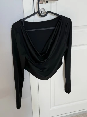 Svart långärmad croppad topp - Snygg svart croppad topp med lång ärm och draperad v-ringning. Perfekt för att styla med höga jeans eller kjol. Tunn och stretchig, vilket gör den bekväm och enkel att bära.
