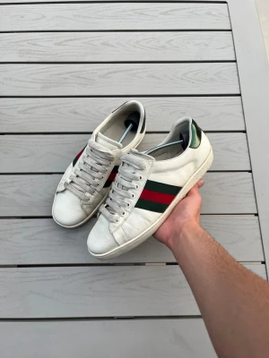vita sneakers med ränder - Sneakers med röda sidoremsor samt metallicgrön detalj på hälen. Låga och stilrena med vita snören och gummisula. Perfekta för dig som vill ha en ikonisk och trendig look.  Priser går att diskuteras 