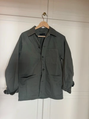 Mörkgrön overshirt från Uniqlo med stora fickor - Stilren mörkgrön overshirt med tre stora utanpåliggande fickor framtill och klassisk krage. Overshirten har knäppning med tre knappar och raka ärmar. Tillverkad i ett slitstarkt bomullstyg som ger en avslappnad look. Storlek S 