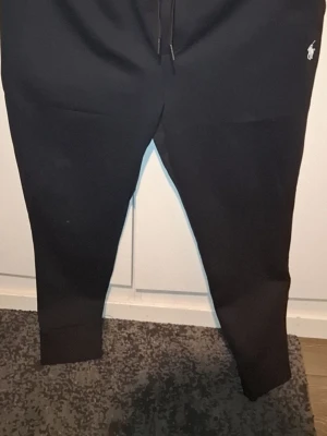 Svart loungewear-set från Polo Ralph Lauren - Mysigt svart loungewear-set från Polo Ralph Lauren med matchande hoodie och byxor. Hoodien har dragkedja, huva med snören och vit logga på bröstet. Byxorna har resår i midjan, snörning och vit logga vid höften. Perfekt för chill dagar hemma.