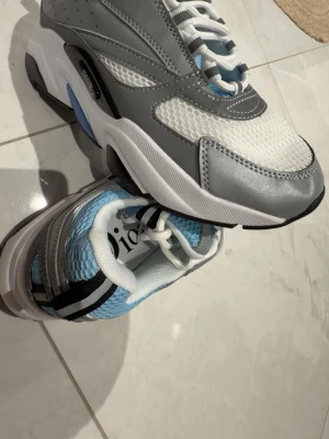 Dior sneakers i grått, vitt och blått -  Dior b22 sneakers med mesh i vitt och blått, samt detaljer i grått och silverfärgat läder. Tjock vit sula med svarta och blå inslag. Klassisk Dior-logga på sidan och bak på hälen. Snygg sportig look med moderna material och färgkombinationer.