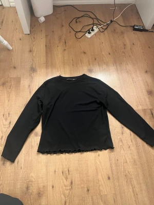 Svart långärmad topp från 157 - En svart långärmad topp från 157 med enkel design och vågig kant nertill. Toppen har rund halsringning och är tillverkad i ett mjukt material som känns skönt mot huden. Den är aldrig använd. Ganska stor i storleken.