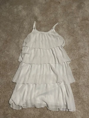 Vit volangklänning från KappAhl - Säljer en söt vit klänning från KappAhl med flera lager volanger och tunna axelband. Klänningen har ett subtilt randigt mönster och är tillverkad i ett lätt, luftigt material som känns skönt mot huden. Perfekt för sommarens alla tillfällen!