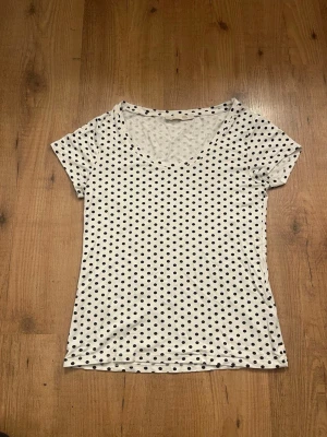 Vit prickig t-shirt i bomull - Snygg vit t-shirt med svarta prickar, perfekt för en avslappnad stil. T-shirten har korta ärmar och är gjord i mjuk bomull som känns skön mot huden. Passformen är normal och den är lätt att matcha med jeans eller kjol.