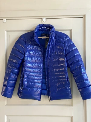 Blå dunjacka från Polo Ralph Lauren - Snygg blå dunjacka från Polo Ralph Lauren med glansig finish och quiltad design. Jackan har hög krage, dragkedja framtill och två sidofickor med dragkedja. Perfekt för kalla vinterdagar och riktigt lätt att bära. Royalblue med tillhörande påse helt ny oanvänd smallpetit