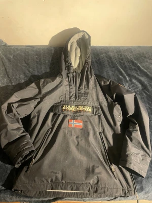 Svart vindjacka från Napapijri - Säljer en svart dunjacka från Napapijri med stor huva och dragkedjor på sidorna. Jackan har en stor ficka framtill med Napapijri-logga och norsk flagga. Perfekt för kalla vinterdagar och riktigt snygg streetstil. Storlek L men hade sagt den passar M bättre. Nypris 2600kr. Vid frågor skicka PM