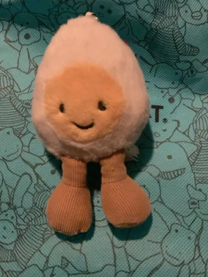 Gosedjur stekt ägg Jellycat - Säljer ett gulligt gosedjur från Jellycat som ser ut som ett stekt ägg med ett leende ansikte. Mjuk plysch i vitt och gult med bruna ben i ribbat tyg. Perfekt som present eller dekoration till rummet.