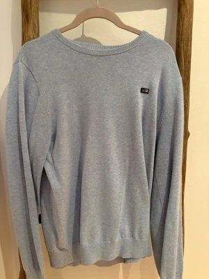 Ljusblå tröja från Lexington - Ljusblå tröja från Lexington med rund halsringning och diskret logotyp på bröstet. Tröjan har långa ärmar och ribbade muddar vid ärmslut och nederkant. Perfekt för en stilren och avslappnad look.