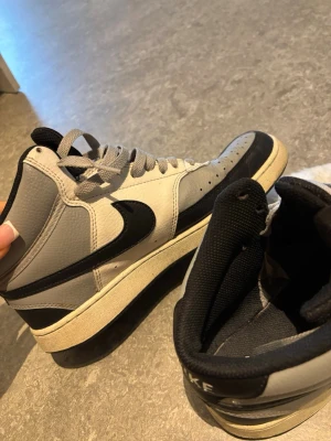 Nike grå och svarta high sneakers - Nike high sneakers i grått och svart med klassisk Swoosh på sidan. Skorna har perforerad tå, vita och grå skosnören samt svart vadderad plös med Nike-logga. Ovandelen är i läder och sulan är vit med svart detalj. Perfekt för dig som gillar retrostil.