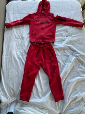Rött Nike set med huva och byxor - Säljer ett rött Nike set med zip hoodie och matchande byxor. Hoodien har svarta detaljer och Nike-logga på bröstet, samt dragkedja framtill. Byxorna har resår i midjan med snörning och Nike-logga på benet. Mjukt och bekvämt material, perfekt för chill eller träning. 