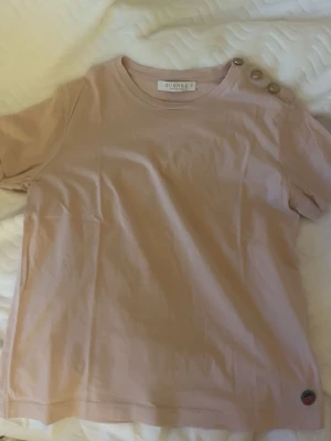 Ljusrosa t-shirt med knappar från Busnel - Snygg ljusrosa t-shirt från Busnel med rund halsringning och tre dekorativa guldknappar på ena axeln. Tillverkad i mjukt bomullstyg och har en diskret Busnel-logga vid nederkanten. Perfekt för en clean och stilren look.