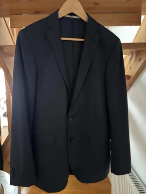 Svart/grå/blå klassisk kostym med knappar - Snygg svart/grå/blå kostym med två knappar framtill och klassiska fickor. Perfekt till mer uppklädda tillfällen. Enkel design med smal krage och diskreta detaljer. Passar dig som gillar stilrena plagg. En liten reva på byxfickan bak se sista bilden.