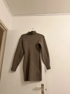 Brun stickad poloklänning från KappAhl - Stickad brun polotröja från KappAhl med lång ärm och ribbad struktur. Tröjan har en hög krage och är något längre i modellen, vilket ger en snygg och avslappnad siluett. Perfekt för lager på lager och höstmys.