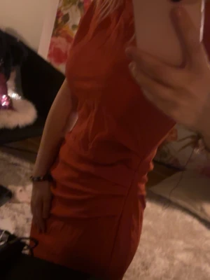 Orange omlottklänning oneshoulder - Snygg orange kortklänning med omlottdetalj och oneshoulder-design. Klänningen har en figurnära passform och är perfekt för dig som vill sticka ut. Materialet känns mjukt och följsamt mot kroppen.