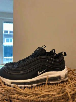 Nike Air Max 97 svarta sneakers - Nike Air Max 97 sneakers i svart med vit sula och synlig Air Max-dämpning. Skorna har vågformade detaljer i textil och syntet, samt en liten vit Swoosh på sidan. Klassisk snörning och dragflikar på plös och häl för enkel påtagning.