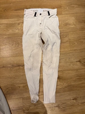 Vita ridbyxor från Kingsland - Säljer ett par vita ridbyxor från Kingsland med förstärkta knäpartier och smal passform. Byxorna har bälteshällor, dubbla knappar i midjan och elastiska benslut. Perfekta för dig som gillar stilrena och funktionella ridkläder.