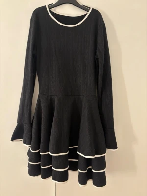 Svart volangklänning med vita kanter - Supersnygg svart klänning med långa ärmar och flera volanger i kjolen. Klänningen har vita kontrastkanter vid halsringning, ärmslut och volanger. Materialet är tjockare och har ett diskret mönster, perfekt för kyligare dagar. Ger en cool och unik look!