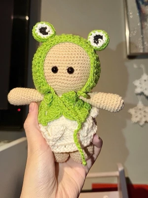 Virkad amigurumi med grodmössa - Supergullig handvirkad docka med beige kropp och svartbroderade ögon. Dockan har en grön mössa med grodögon och en matchande grön sjal. Virkad med bomullsgarn.