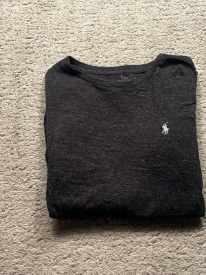 Mörkgrå tröja från Polo Ralph Lauren - Snygg mörkgrå tröja från Polo Ralph Lauren med klassisk vit broderad logga på bröstet. Tröjan har en enkel och stilren design, perfekt till alla tillfällen. Mjuk och bekväm med rund halsringning.